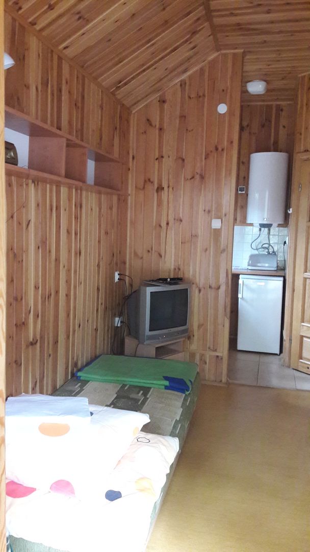 Zdjęcie przedstawia mały domek z lodówką, bojlerem i drzwiami. Z prawej strony kanapa, szafa i stół.